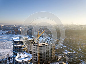 Apartment or office tall building under construction, aerial view. Tower crane silhouette Snowy field and distant city on bright