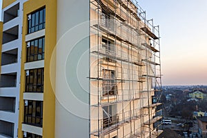 Apartment or office tall unfinished building under construction. Brick wall in scaffolding, shiny windows and tower crane on urban