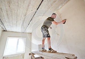 Apartment remodel. Worker is standing on wooden stand and puttying the walls indoors at small room