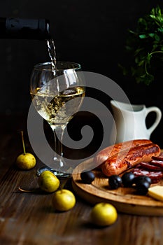 Aperitif concept. Wine pours into a glass goblet. On the table, meat appetizer, fried sausages, salami, cheese, olives.