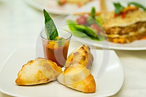 Apetizer of small tomato soup in shotglass and delicious mini empanadas spread around on white plate