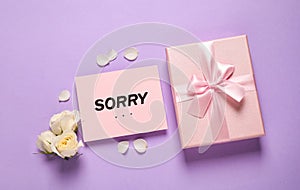 Apology. Pink card with word Sorry, gift box and beautiful roses on violet background, flat lay