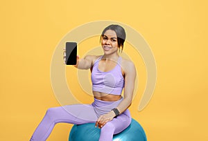 App for workout. Happy black lady holding cellphone with black creen, sitting on fitball over yellow background