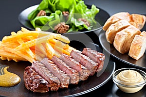 Appetising steak slices served with fries , green salad and baguette on a black plate.