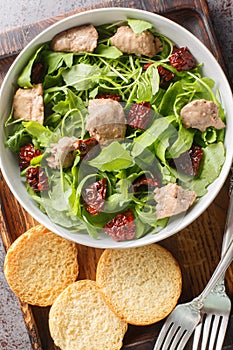 Appetizer balanced with canned cod liver, fresh arugula and sun-dried tomatoes served with toast close-up in a bowl on the table.