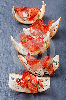 Appetizer bruschetta with jerky prosciutto on thinly sliced ciabatta bread on stone slate background close up.