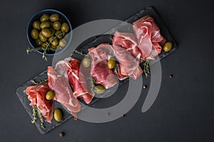 Appetizer from dry cured serrano ham or Spanish jamon iberico. Italian prosciutto crudo served with green olives on the black