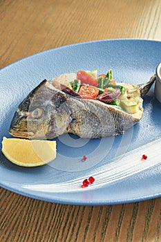 Appetizing baked dorado fish with vegetables tomatoes, asparagus, avocado with yellow sauce on a blue plate on a wooden