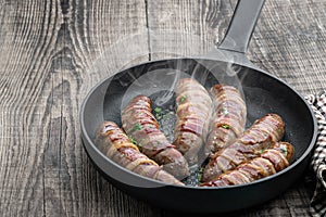 Appetizing sausages wrapped in bacon and grilled in frying pan on wooden