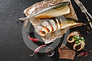 Appetizing smoked fish with spices, cutlery, pepper and bread on craft paper over dark stone background. Sandwich with smoked