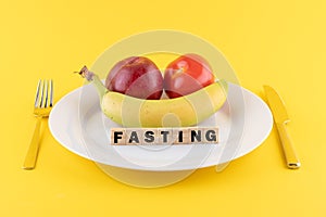 Apple, a banana and a tomato on a plate and the word 'fasting' written on wooden blocks