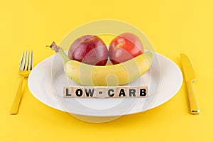 Apple, a banana and a tomato on a plate and the word 'low-carb' written on wooden blocks