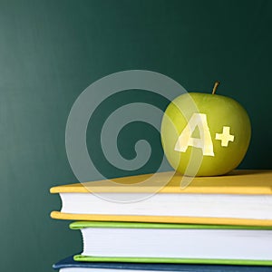 Apple with carved letter A and plus symbol as school grade on books near green chalkboard, space for text