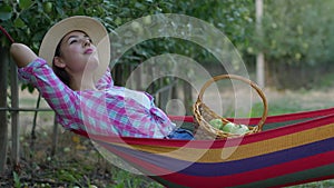 Apple crop, farmer girl dreams while relaxing into hammock and eats fruit from basket after picking up harvest in