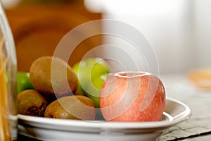 Apple, kiwi and monkey apple are put on white plate over the table in the kitchen with chair as background