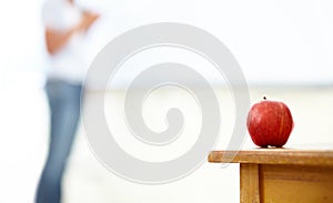 Apple for the teach. An apple on a desk with a woman blurred in the background.