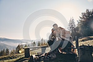 Appreciating every minute together. Beautiful young couple having morning coffee while sitting by the campfire in mountains