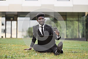 African businessman sitting on green grass outside office, showing thumb up while using laptop