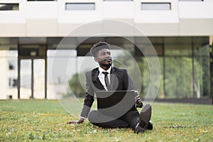 African businessman sitting on green grass outside office, showing thumb up while using laptop