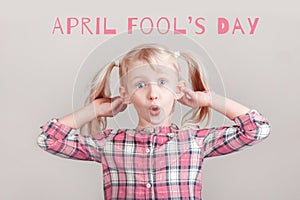 April Fools Day card with text. Funny Caucasian girl with pigtails making silly face on light background. Child holding pulling