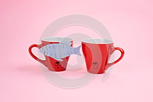 April Fools ' Day celebration, paper fish prank. Two joking laughing coffee cups with paper fish on pink background. All Fools ' D