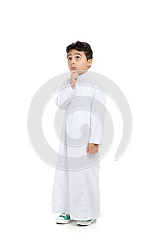 Arab boy with a confused look and his hand on chin, looking up, wearing white traditional Saudi Thobe and sneakers, raising his photo