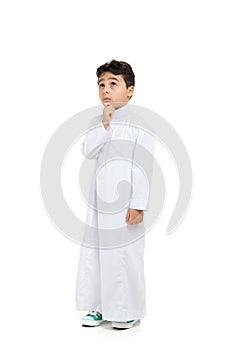 Arab boy with a confused look and his hand on chin, looking up, wearing white traditional Saudi Thobe and sneakers, raising his