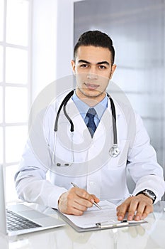 Arab doctor man using laptop computer while filling up medication history records form at the glass desk in medical
