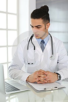 Arab doctor man using laptop computer while filling up medication history records form at the glass desk in medical