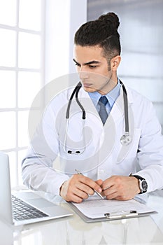 Arab doctor man using laptop computer while filling up medication history records form at the glass desk in medical