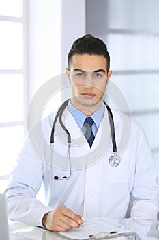 Arab doctor man using laptop computer while filling up medication history records form at the glass desk in medical