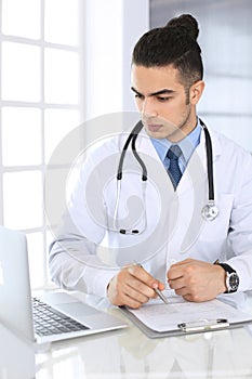 Arab doctor man using laptop computer while filling up medication history records form at the glass desk in medical