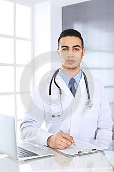 Arab doctor man using laptop computer while filling up medication history records form at the glass desk in medical