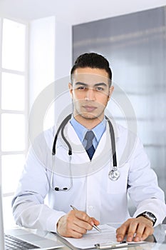 Arab doctor man using laptop computer while filling up medication history records form at the glass desk in medical