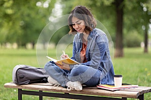 Arab female student writing in notebook while sitting on bench in park