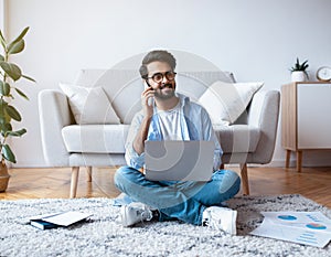 Arab Freelancer Guy Using Laptop And Talking On Cellphone At Home