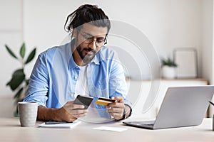 Arab freelancer guy using smartphone and credit card at home, paying online