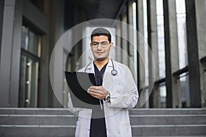 Arab Indian male doctor, standing outside modern clinic, making notes and filling in patient's record