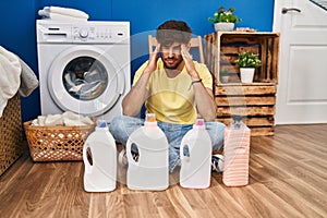 Arab man with beard doing laundry sitting on the floor with detergent bottle with hand on head, headache because stress