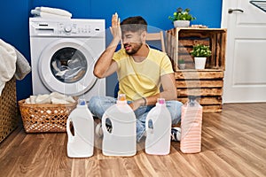 Arab man with beard doing laundry sitting on the floor with detergent bottle surprised with hand on head for mistake, remember