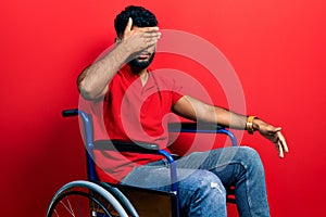 Arab man with beard sitting on wheelchair covering eyes with hand, looking serious and sad