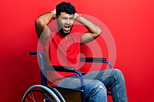 Arab man with beard sitting on wheelchair crazy and scared with hands on head, afraid and surprised of shock with open mouth