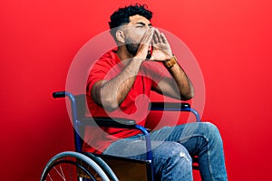 Arab man with beard sitting on wheelchair shouting angry out loud with hands over mouth