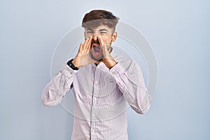 Arab man with beard standing over blue background shouting angry out loud with hands over mouth