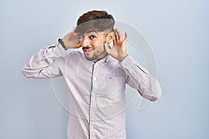 Arab man with beard standing over blue background trying to hear both hands on ear gesture, curious for gossip