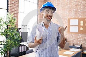 Arab man with beard wearing architect hardhat at construction office pointing thumb up to the side smiling happy with open mouth