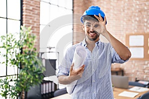 Arab man with beard wearing architect hardhat at construction office smiling happy doing ok sign with hand on eye looking through