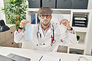 Arab man with beard wearing doctor uniform holding medical mask relaxed with serious expression on face