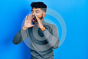 Arab man with beard wearing turtleneck sweater shouting angry out loud with hands over mouth