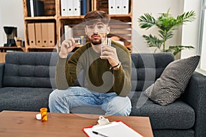 Arab man with beard working on depression holding pills and water relaxed with serious expression on face